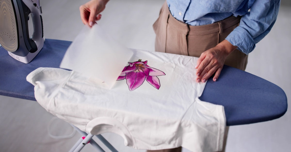 A person removes a sheet from the top of a pink floral design centered on a white T-shirt. An iron is nearby.