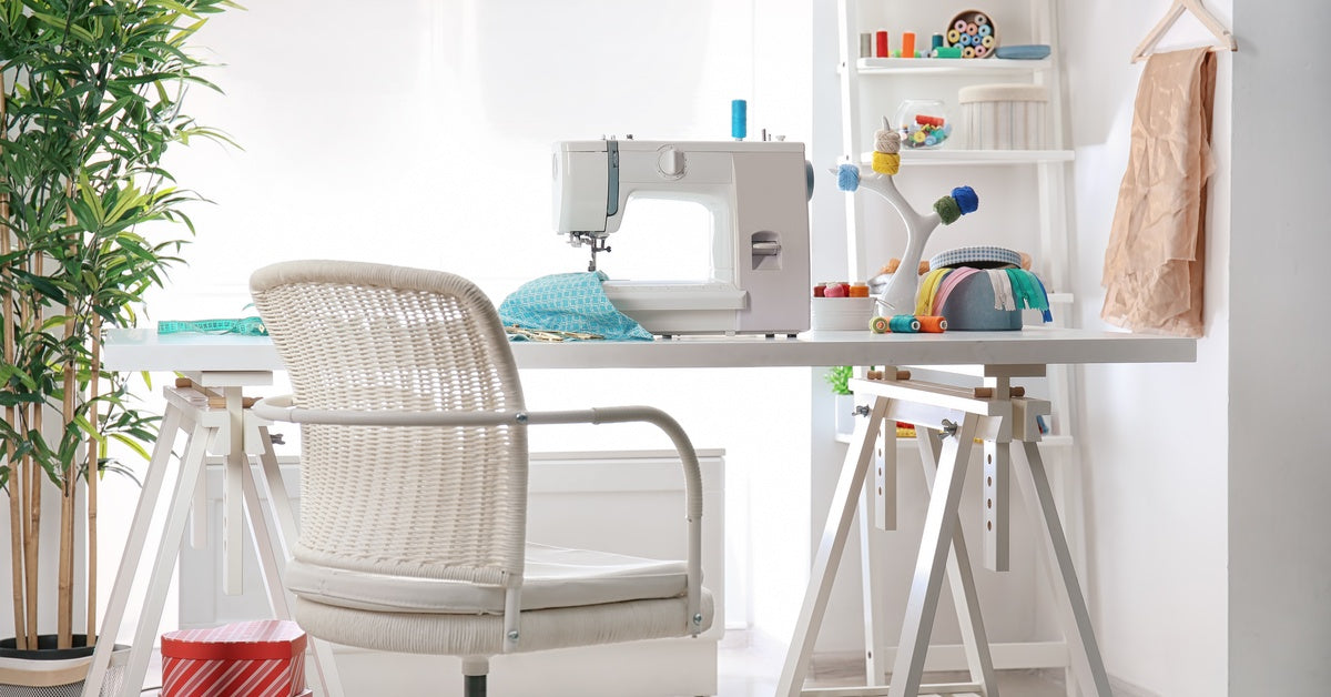 A bright sewing workspace features a table on sawhorse-style legs, a white swivel chair, and ladder shelves.
