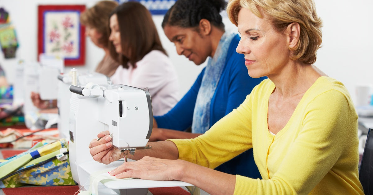 Several people sitting in a row work at sewing machines in a classroom setting. Textile pieces hang on the wall.
