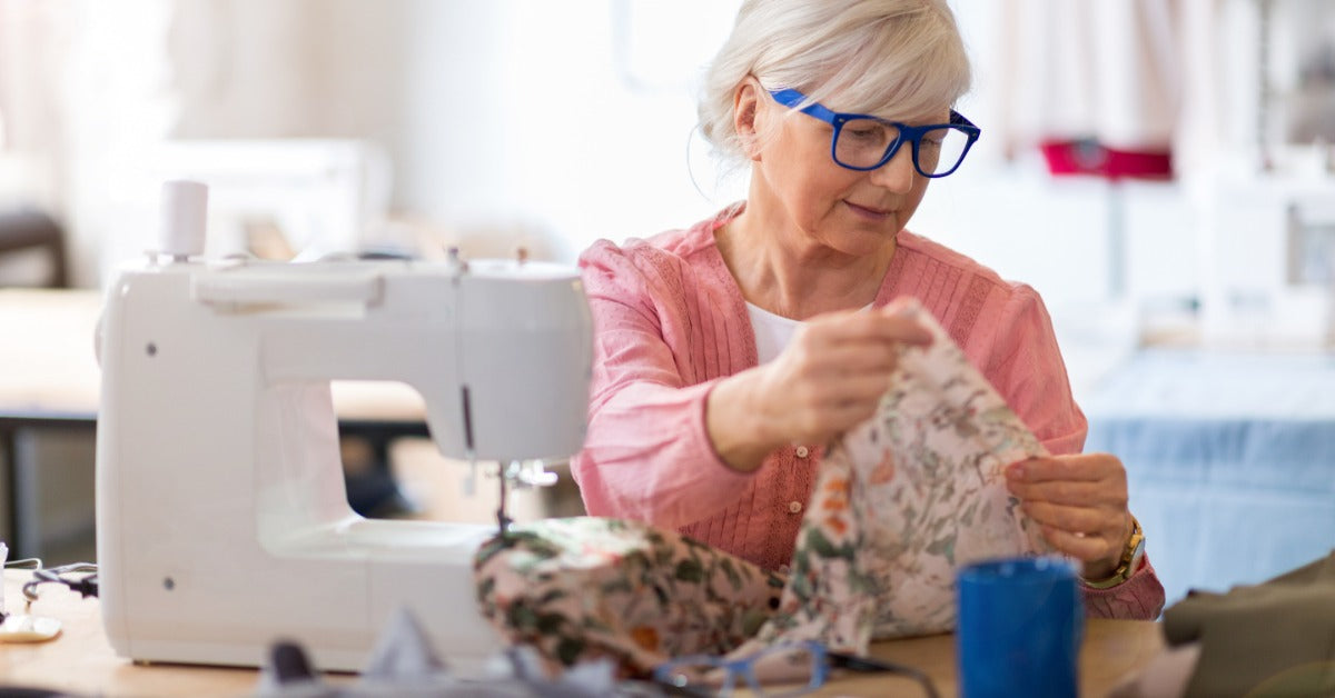 An older woman in blue glasses sits at a sewing machine. She examines patterned fabric that she holds with both hands.