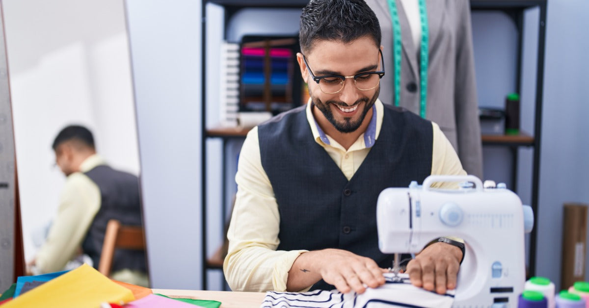 A smiling man in glasses sews striped fabric on a sewing machine. Behind him are shelves and a jacket on a dress form.