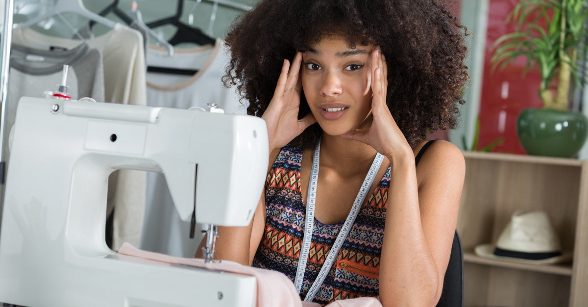 A woman at a sewing table looks at the viewer with eyebrows slightly raised. Her fingertips touch the sides of her face.