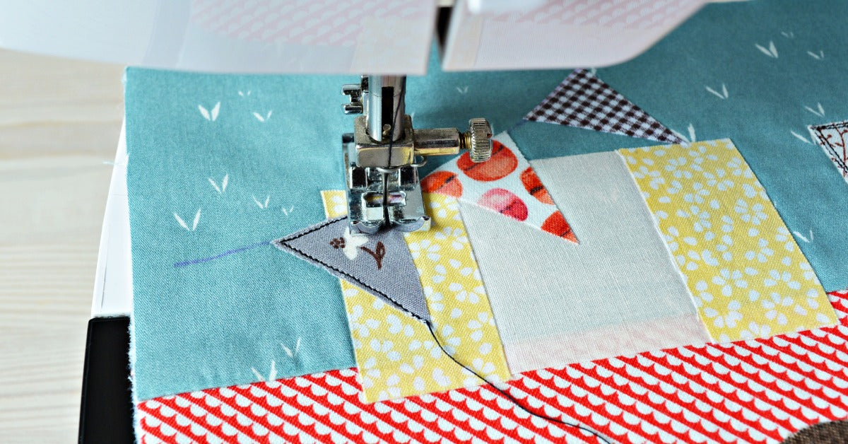 A sewing machine stitches on a colorful quilt block. The presser foot is on top of a small triangular piece of fabric.