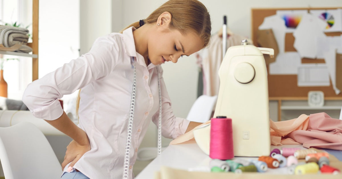 A woman sits at a sewing machine. Her head hangs down as she leans forward and presses her hand to her lower back.