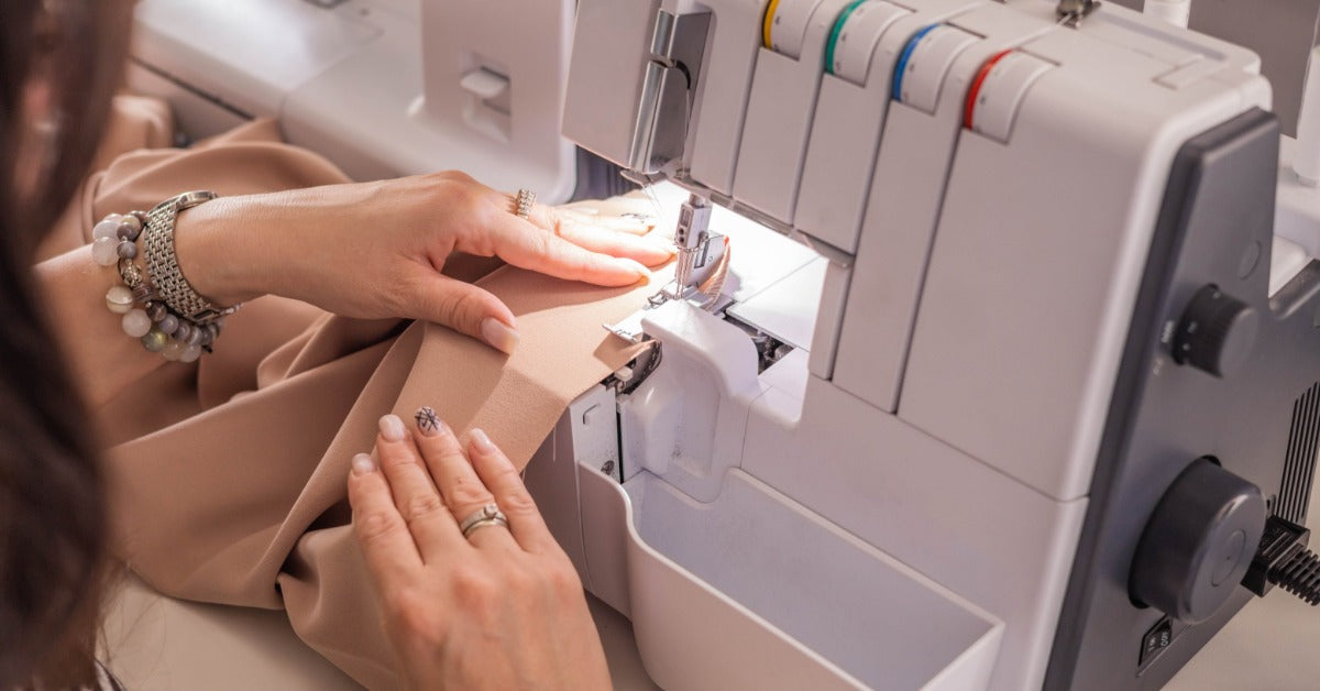 A person guides fabric beneath a serger's presser foot. The machine's front has colored thread guides and control dials.