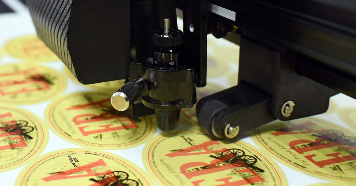 An electronic cutting machine works on a sheet of round yellow labels with red lettering and illustrated details.