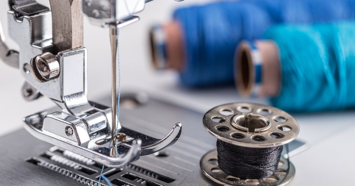 A close-up of a sewing machine presser foot and needle threaded with blue thread. A metal bobbin is on the machine bed.