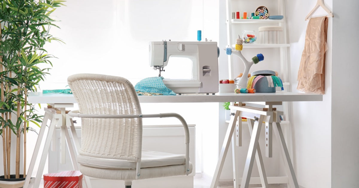 A room features a worktable, a sewing machine, and a cushioned white chair. Colorful thread spools sit on shelves.