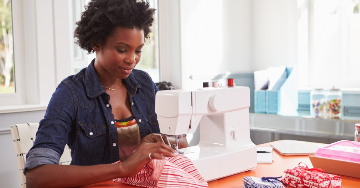 A woman uses a sewing machine on an orange table near a window. She guides striped fabric beneath the needle.