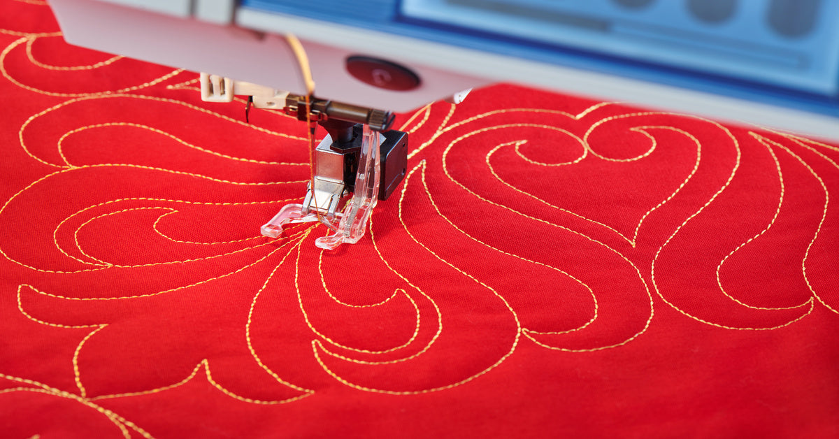 Close-up of a quilting sewing machine needle stitching a pattern in golden thread on bright red fabric.