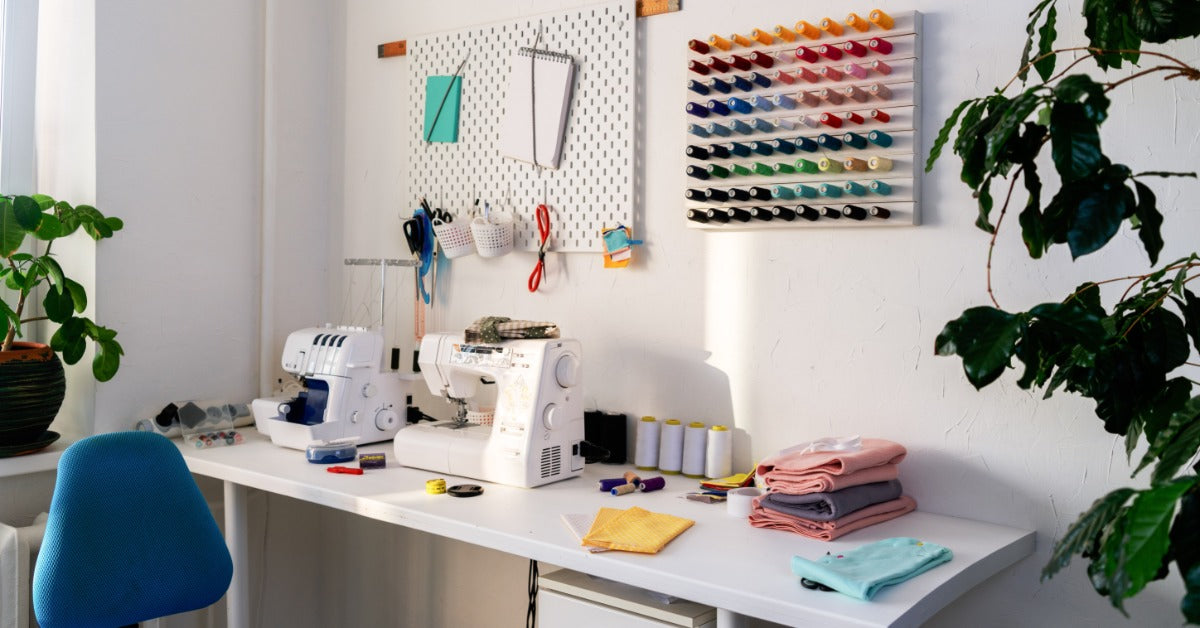 A sewing machine and an overlock machine are on a table. Fabric and spools of thread in different colors are nearby.