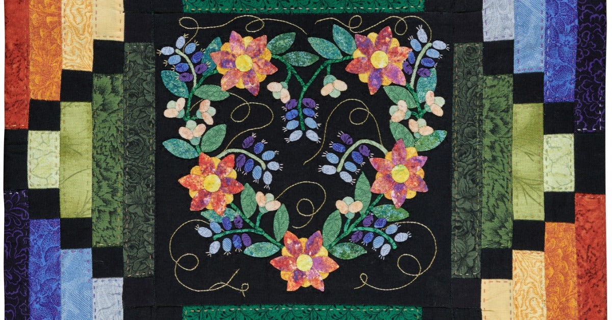 A quilt features a floral appliqué in the center of a black panel. Colorful rectangular fabric pieces frame the center.