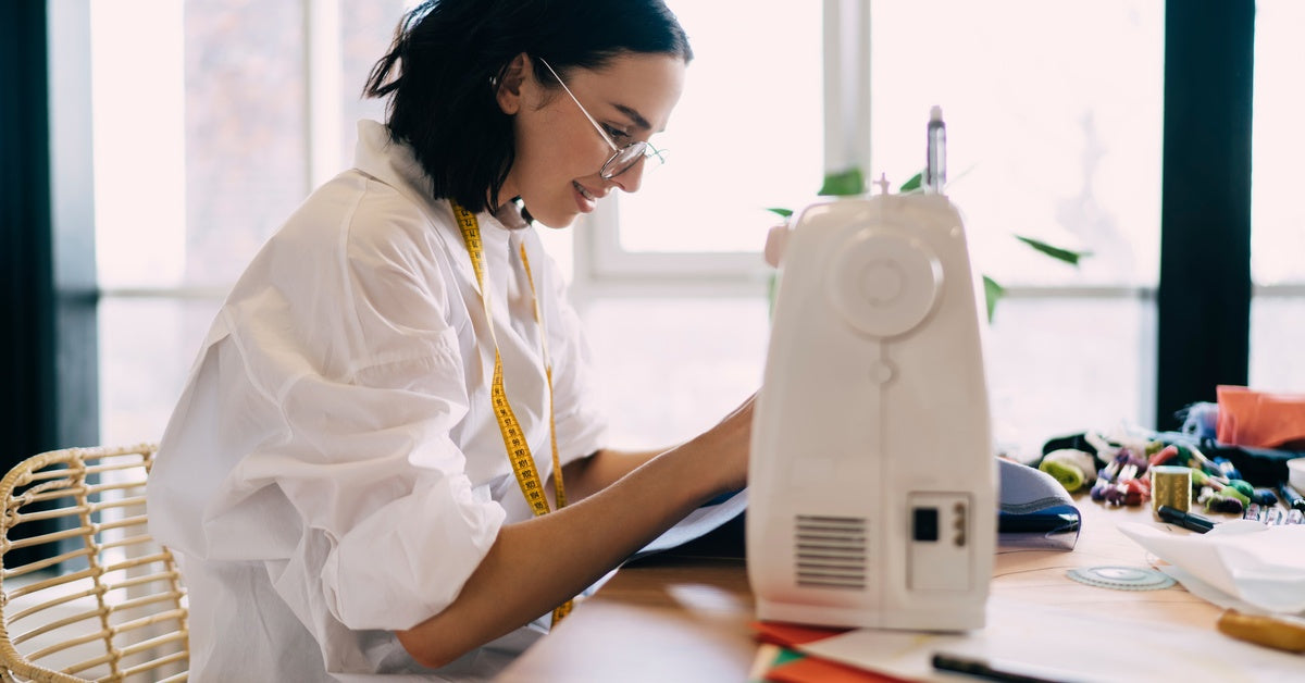 A woman with a measuring tape draped around her neck uses a sewing machine. Fabric and sewing tools are on the tabletop.