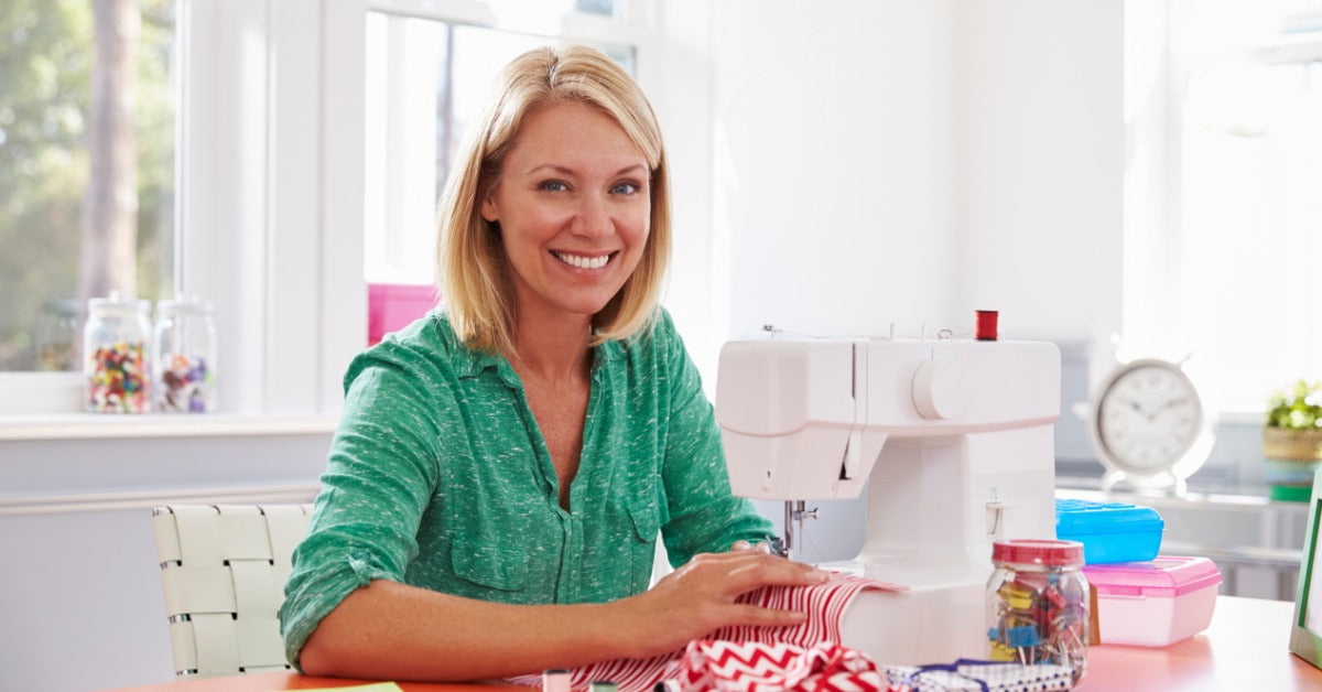 A woman sits at a table in a bright white room. She holds red-and-white striped fabric under a sewing machine needle.