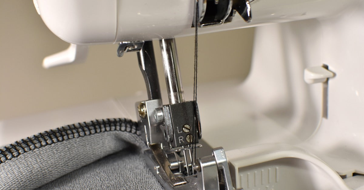 The needles of a white overlock machine sew an overlock stitch as the presser foot guides the gray fabric.