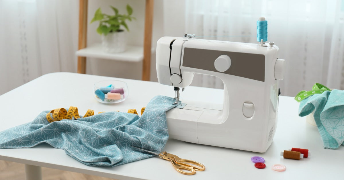 A white sewing machine and other supplies sit on a white table. Blue patterned fabric is gathered under the needle.