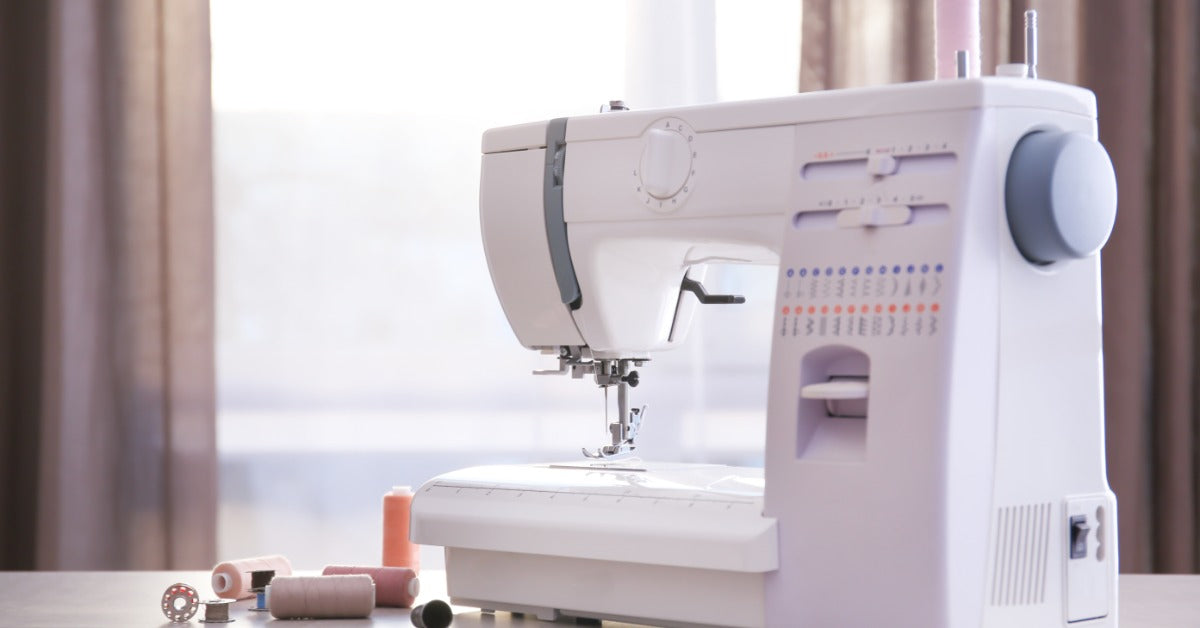 A sewing machine sits beside thread spools and a bobbin on a tabletop. Light shines through a window behind the machine.