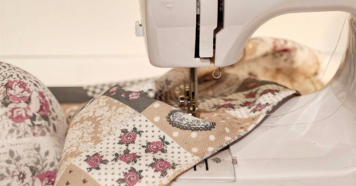 A sewing machine stitches through a quilt in beige, gray, and rose prints. A stitch guide is at the top of the machine.