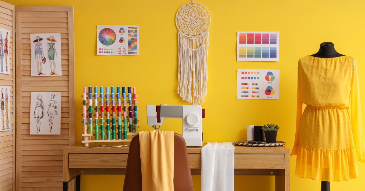 A sewing machine and colorful thread spools are on a wooden table in a yellow room. A yellow dress is on a dress form.