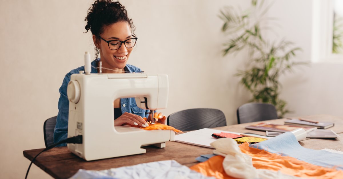A woman in glasses is sitting at her sewing machine, sewing an orange square of fabric. There are various fabrics on the table.